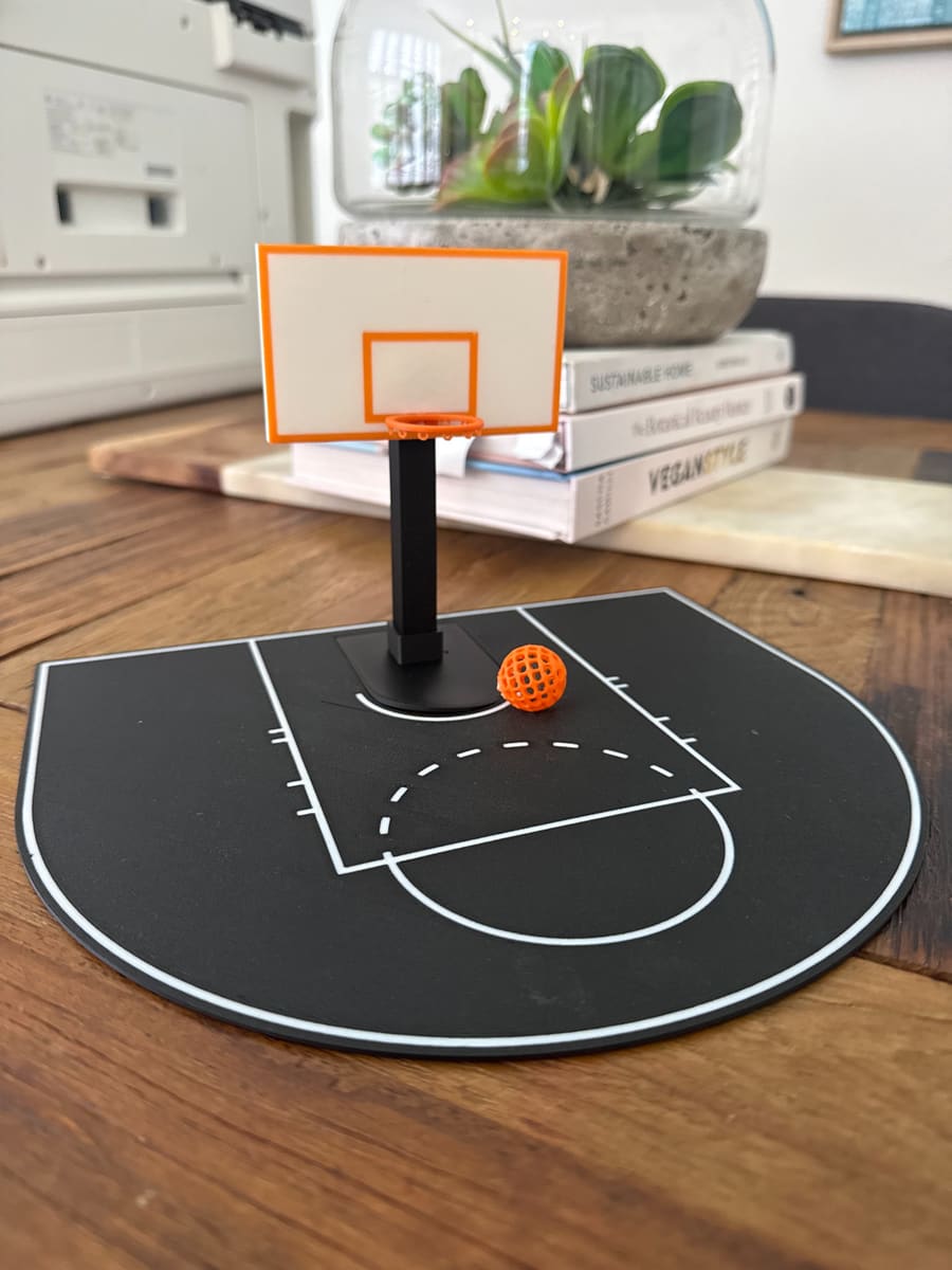 A 3D-printed mini basketball court with a hoop, backboard and an orange lattice ball on a timber table