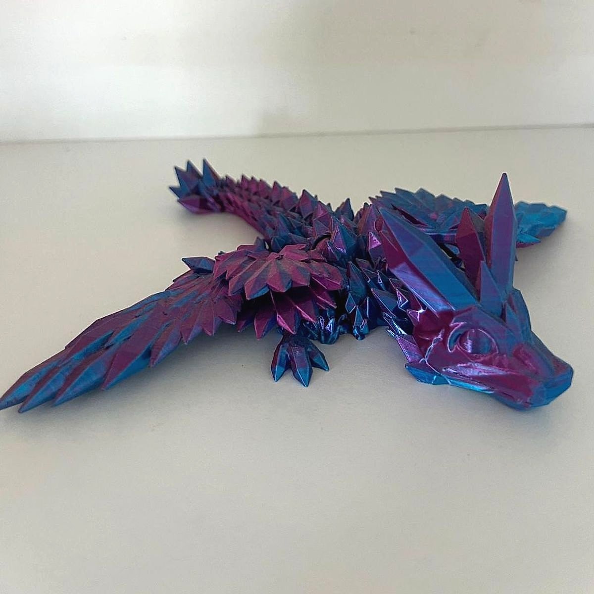 A purple and blue articulated crystal dragon with its wings spread out on a white shelf
