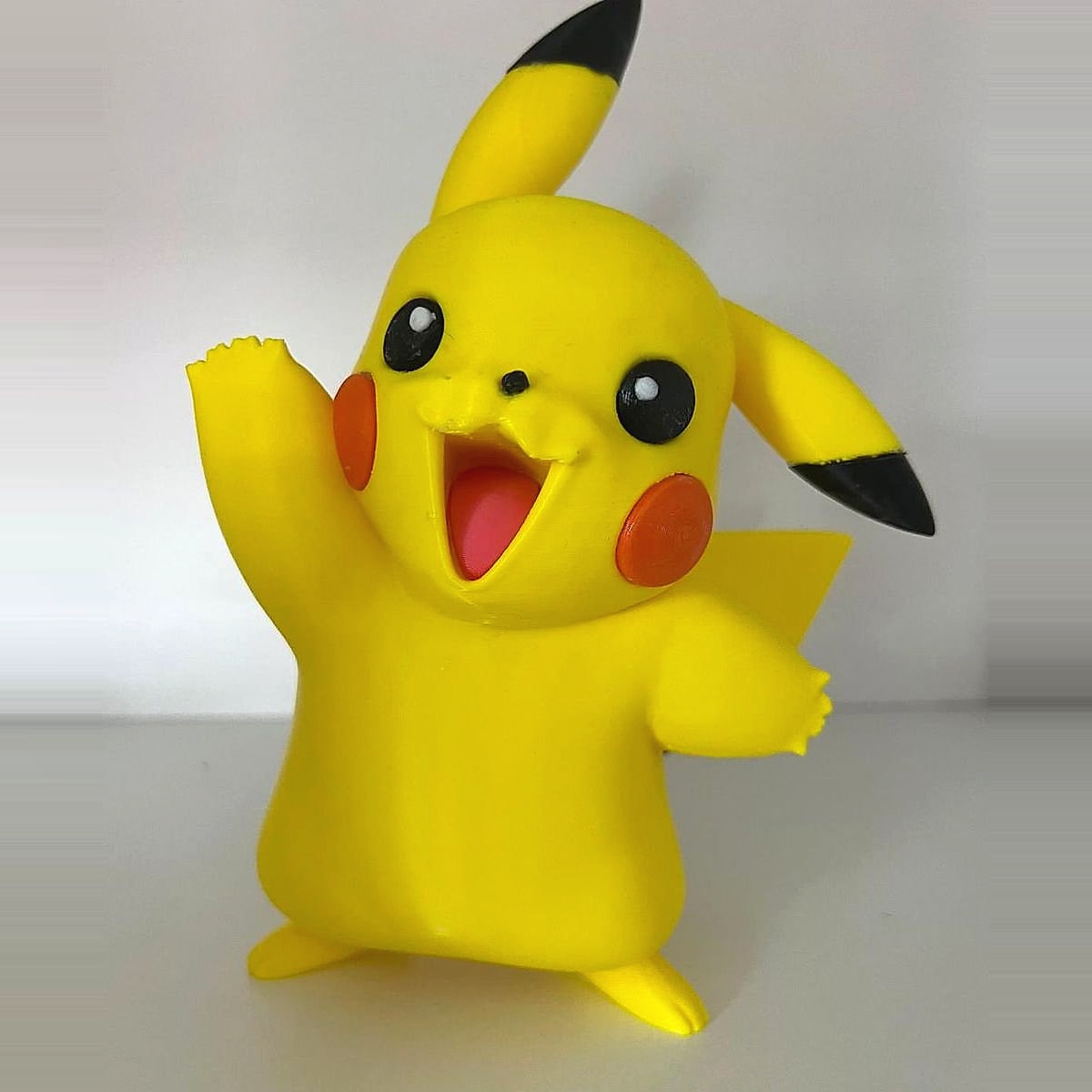 A yellow 3D-printed figurine with red cheeks and black-tipped ears, one arm waving