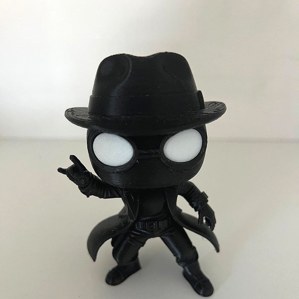 A small black 3D-printed figure wearing a fedora and long coat with big white eyes and one hand raised